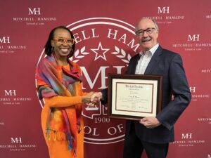Frank Harris '75 receives a handshake and certificate of trustee emeritus status from President and Dean Camille M. Davidson in front of a Mitchell Hamline backdrop.